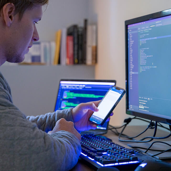 A programmer uses a laptop, computer, and smartphone to work on code for a web software development project.